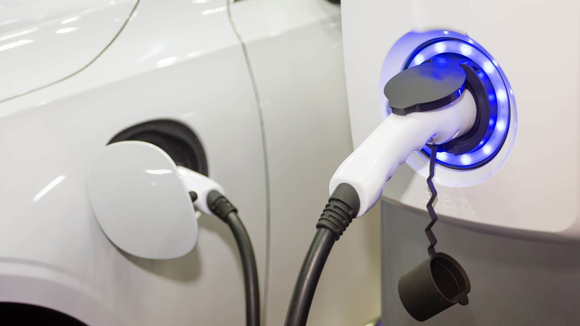 White electric car charging with a cable plugged into a charging station, which has blue lights.