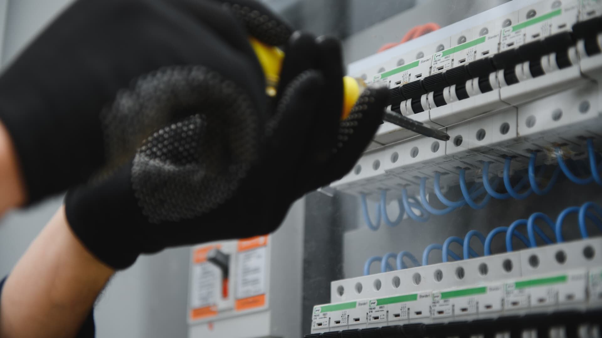 Person wearing black gloves using a screwdriver on a fuse box with blue wires.