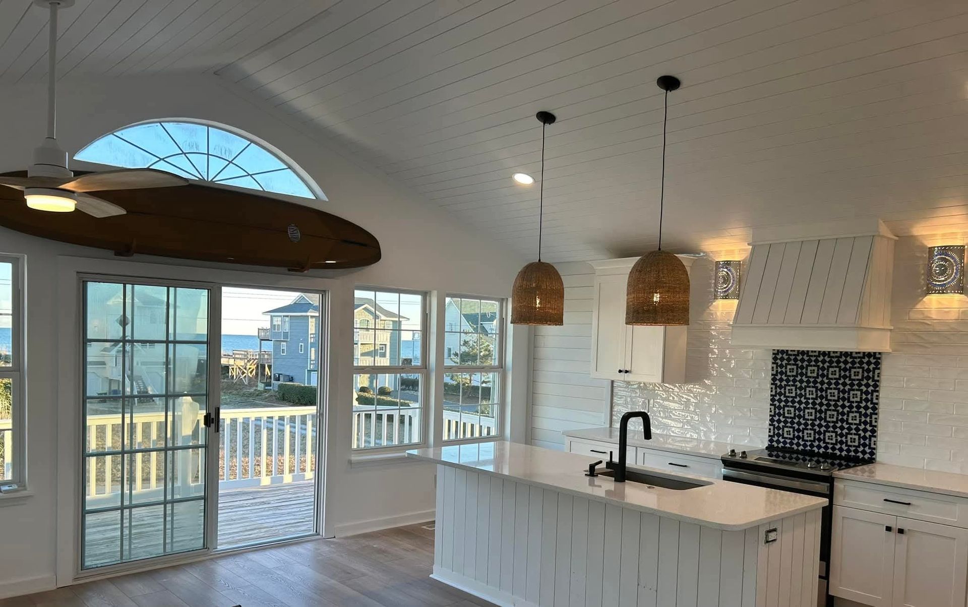 Open-concept kitchen with white cabinetry, island, and high ceiling, featuring a hanging surfboard and view of the sea.