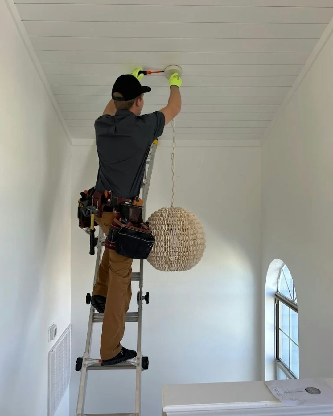 Electrician on a ladder installing a ceiling light fixture in a white room.