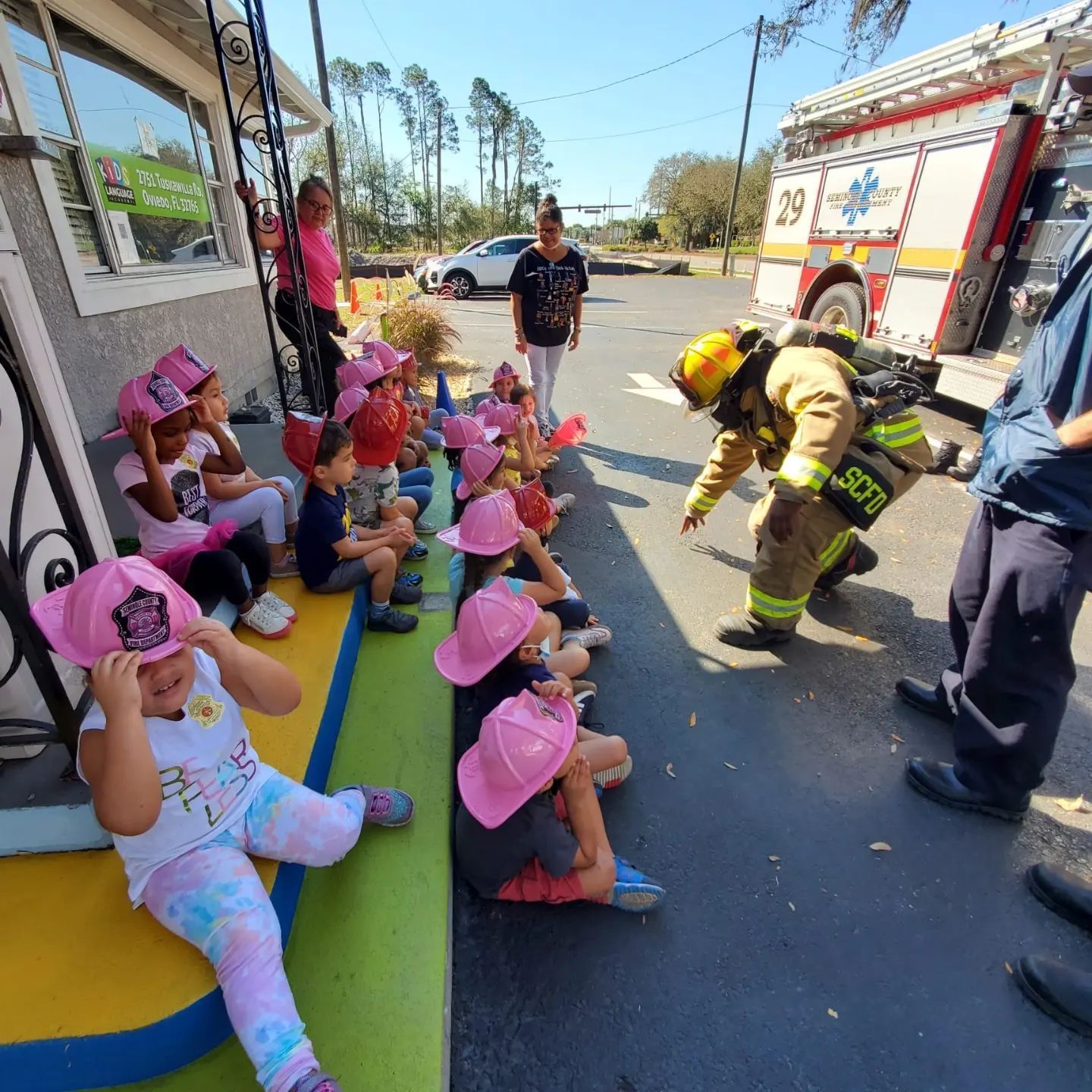 A group of children wearing pink hats are sitting in front of a fire truck