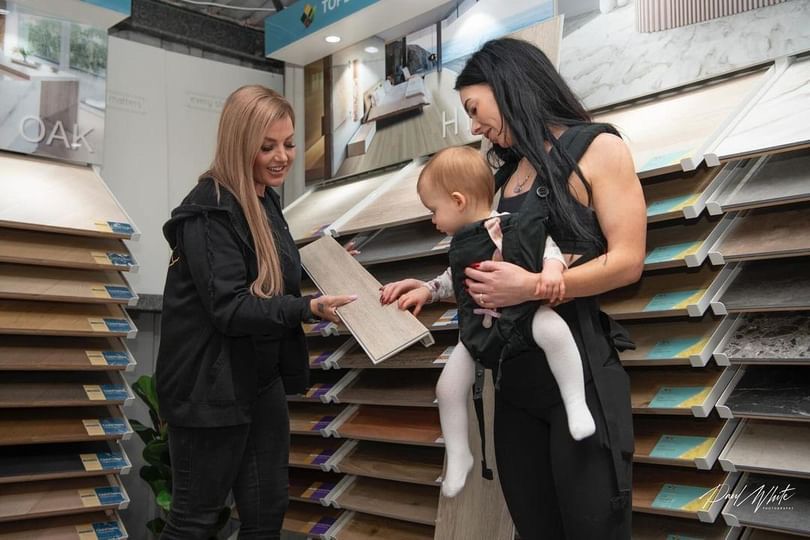A woman shows flooring samples to a customer holding a baby in a showroom.
