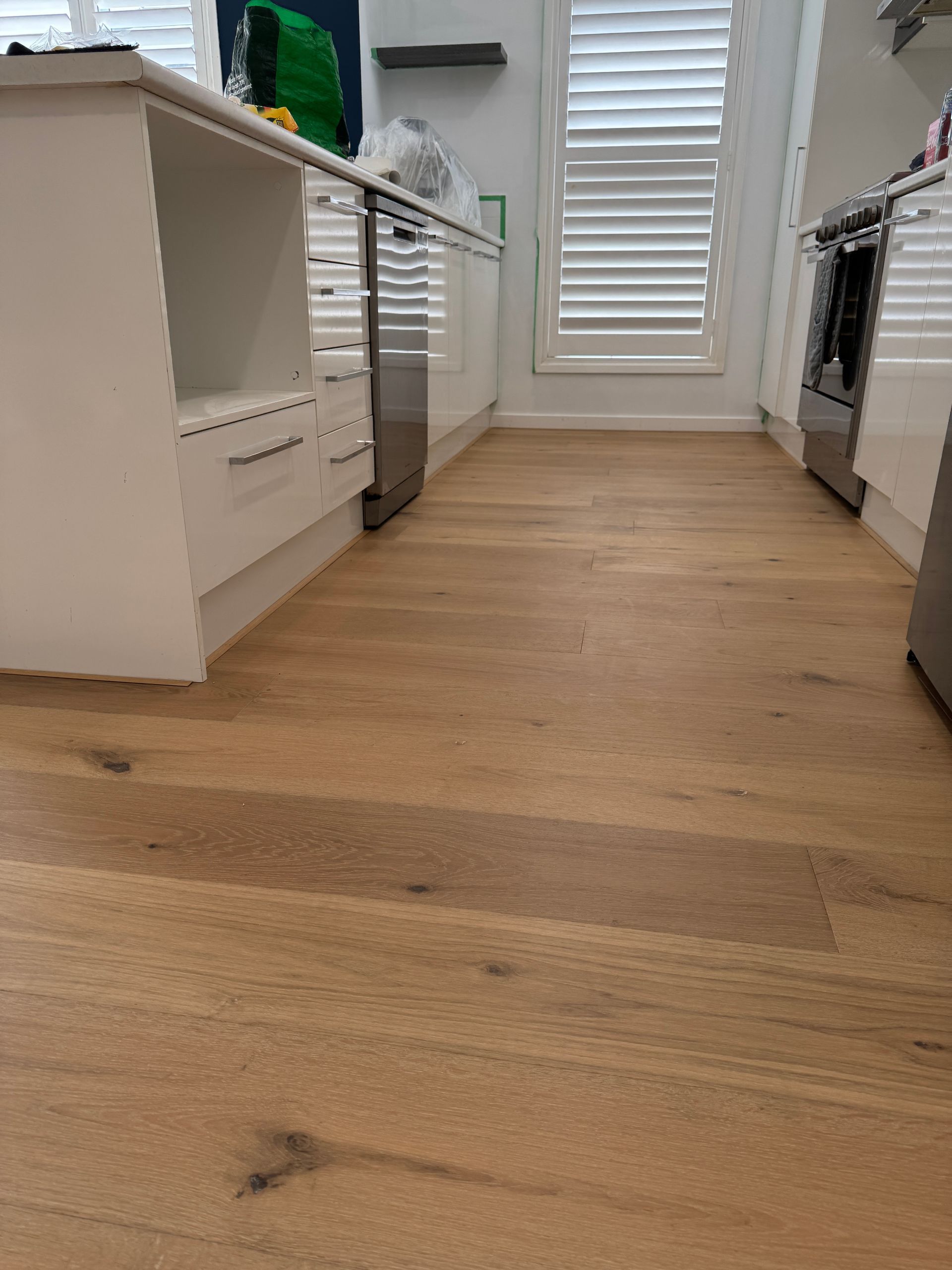 Hardwood floors in a light-filled room, leading to a kitchen with white cabinets, and a hallway with artwork.