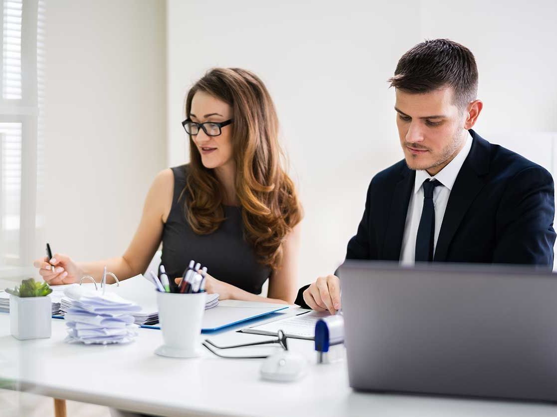 To kontoransatte ved et skrivebord; kvinde med briller skriver, mand i jakkesæt arbejder på en bærbar computer.