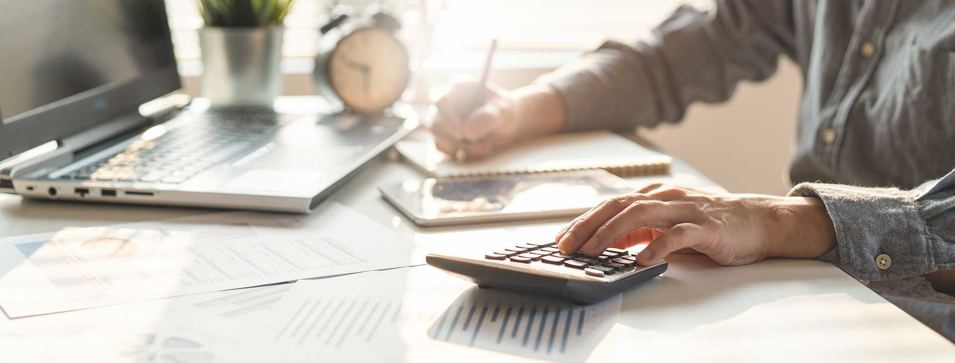Person, der beregner med lommeregner, mens de arbejder på andre dokumenter og en bærbar computer.