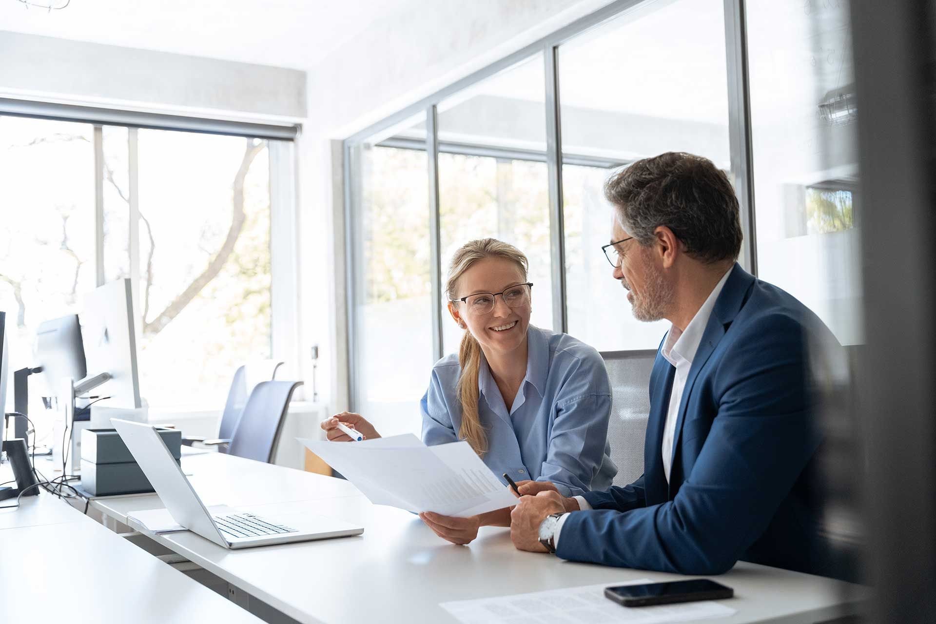 To mænd i et klart oplyst kontor gennemgår dokumenter; den ene gestikulerer, begge ser fokuserede ud.