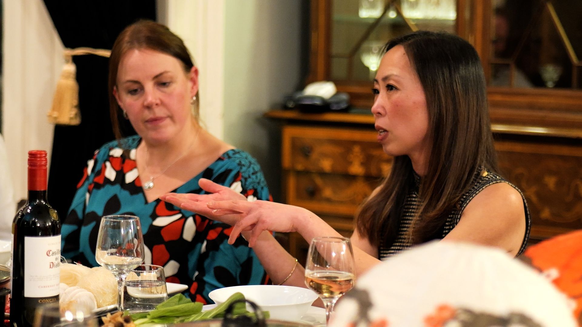 Two women are sitting at a table with wine glasses and a bottle of wine.