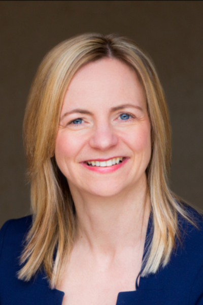 A woman with blonde hair is wearing a blue jacket and smiling.