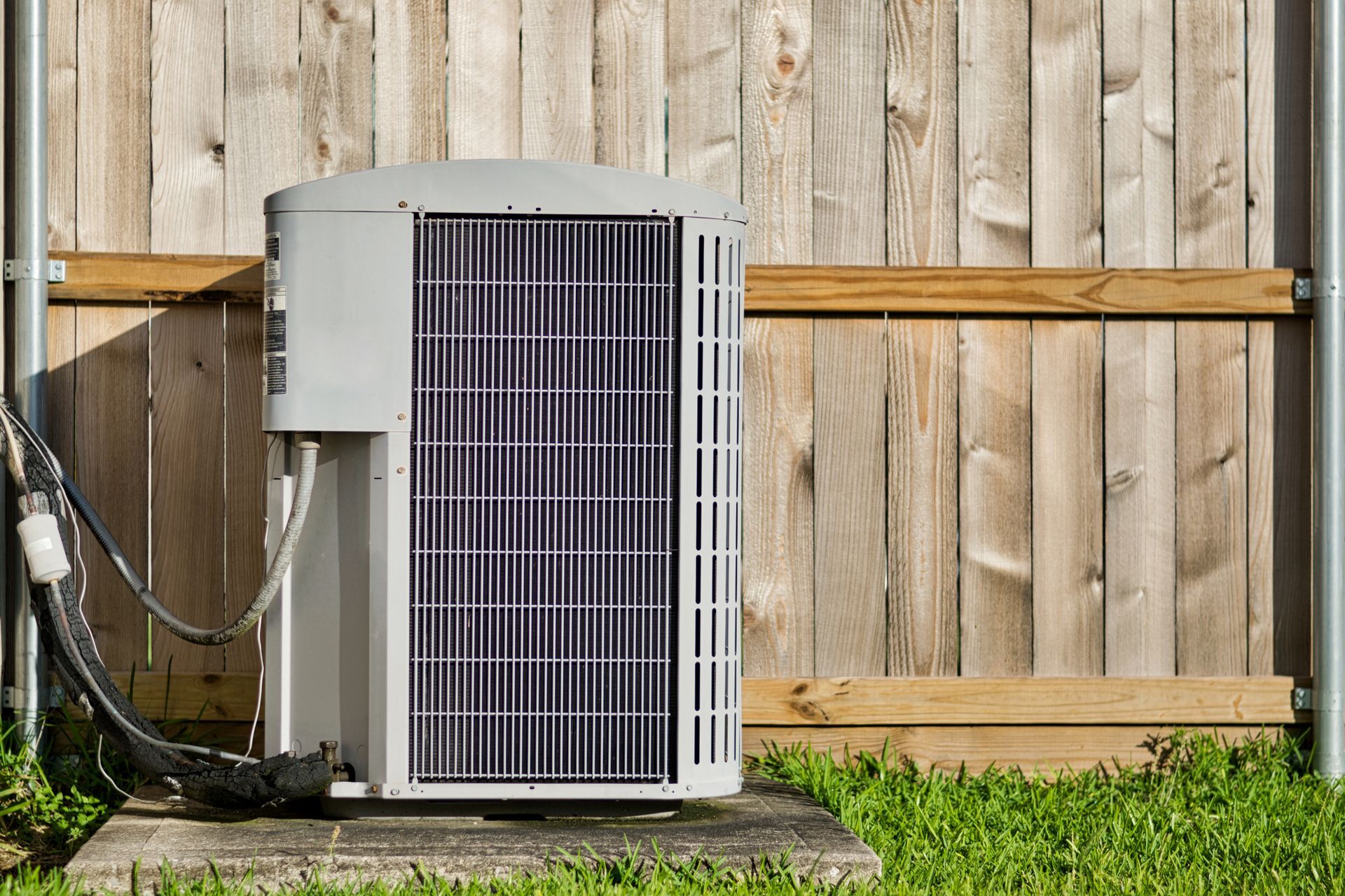Outdoor air conditioning unit against a wooden fence, on a concrete pad, with green grass.