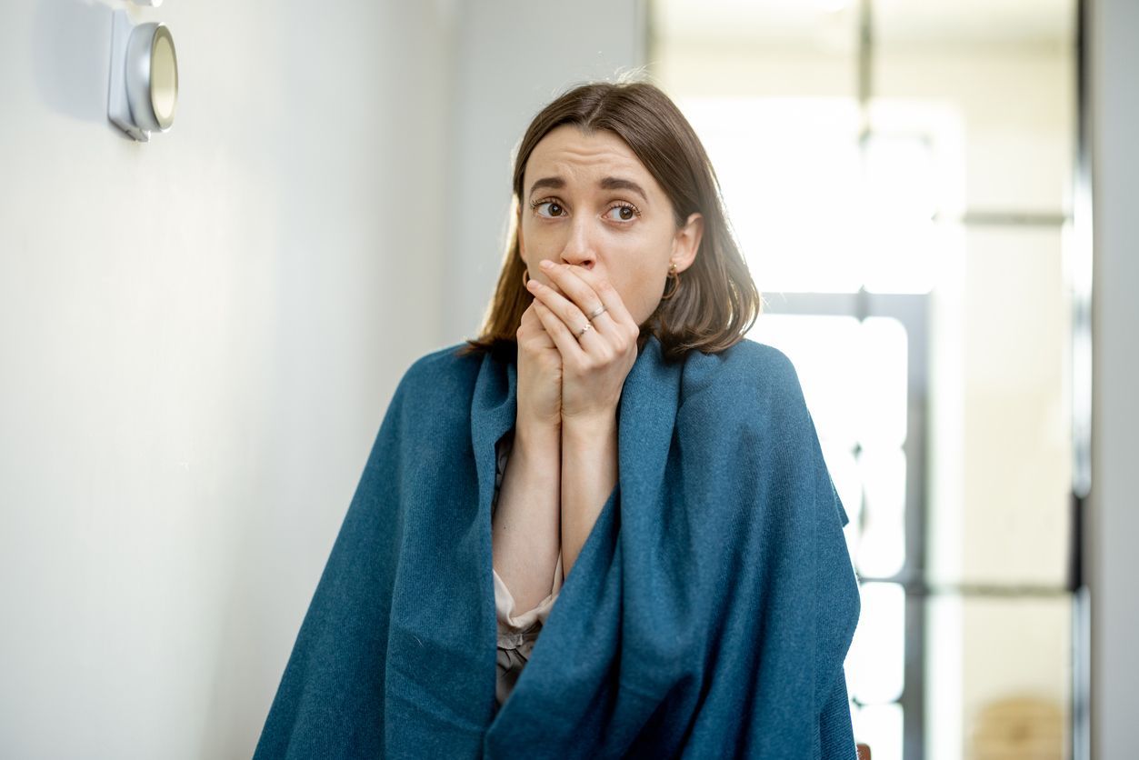 Woman wrapped in blue blanket, looking cold, covers mouth, standing near a thermostat.