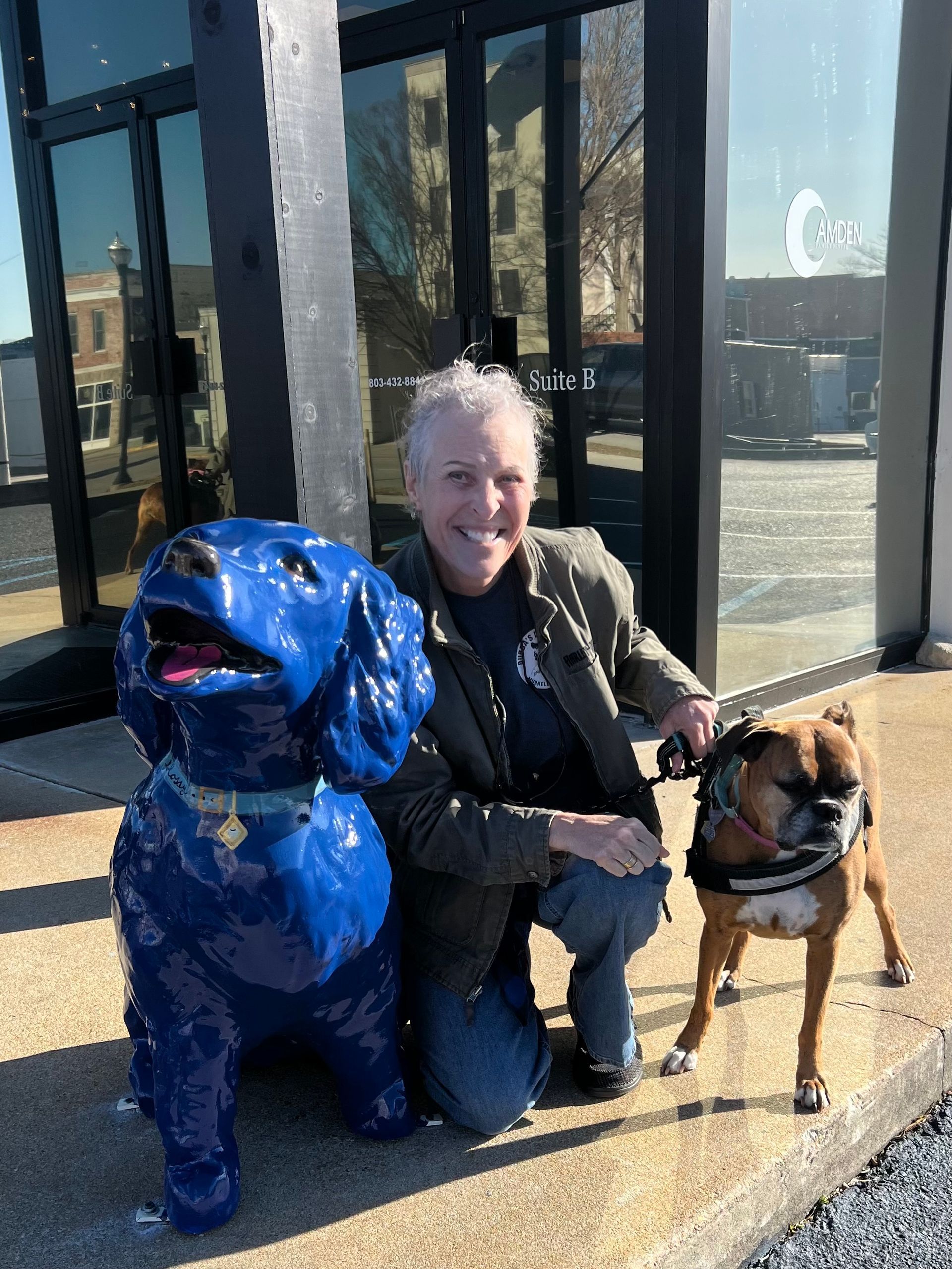 A woman kneels beside a brown dog and a blue dog statue in front of a building.