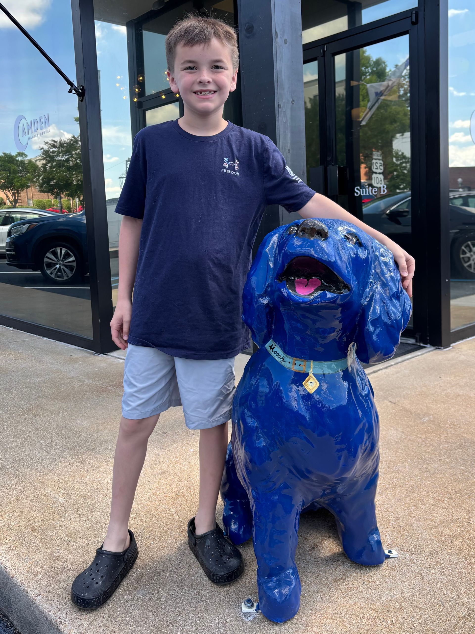 Boy in shorts and shirt smiles next to a large, blue dog statue outside a shop.
