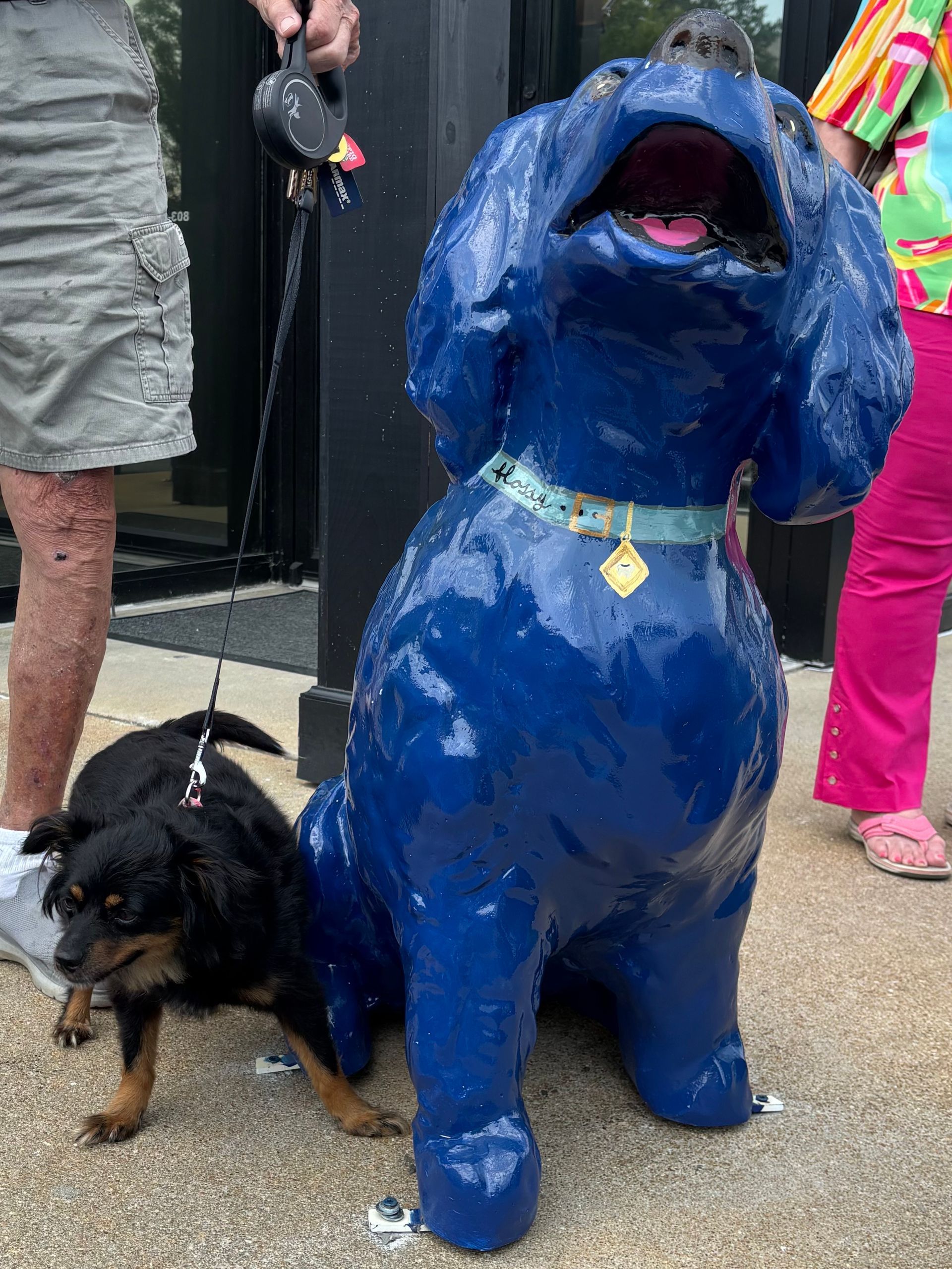 A small black dog next to a large blue dog statue; person holding both leashes. Outdoors.