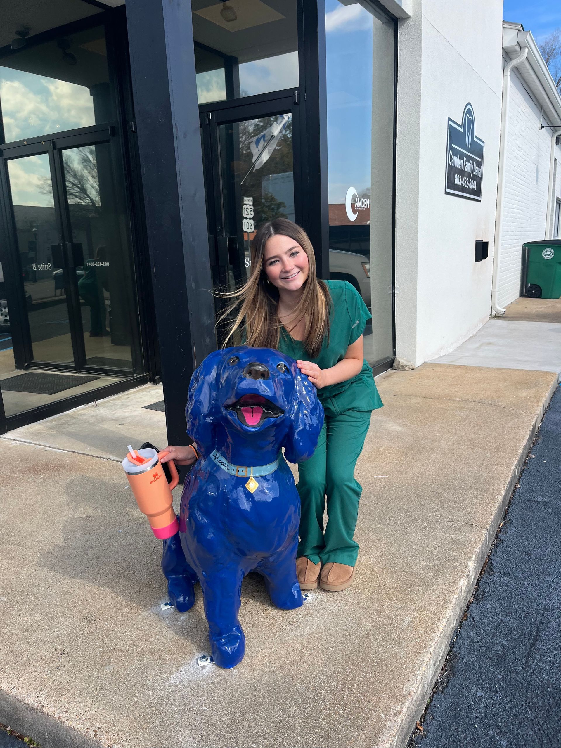 Woman in green scrubs smiles next to a blue dog statue holding a toy outside a building.