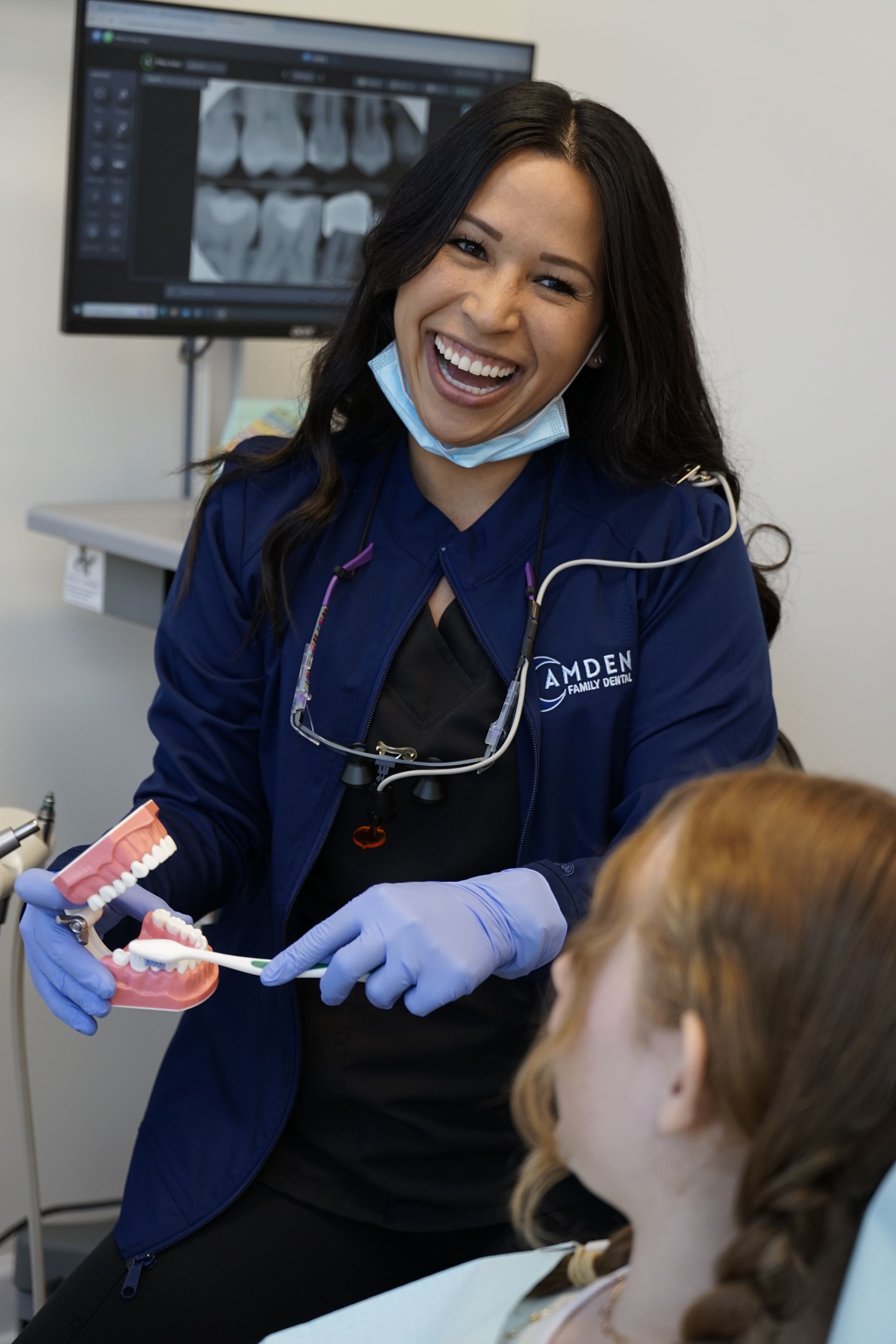 Smiling dentist in blue scrubs, holding teeth model, teaching patient proper brushing technique. Office setting.
