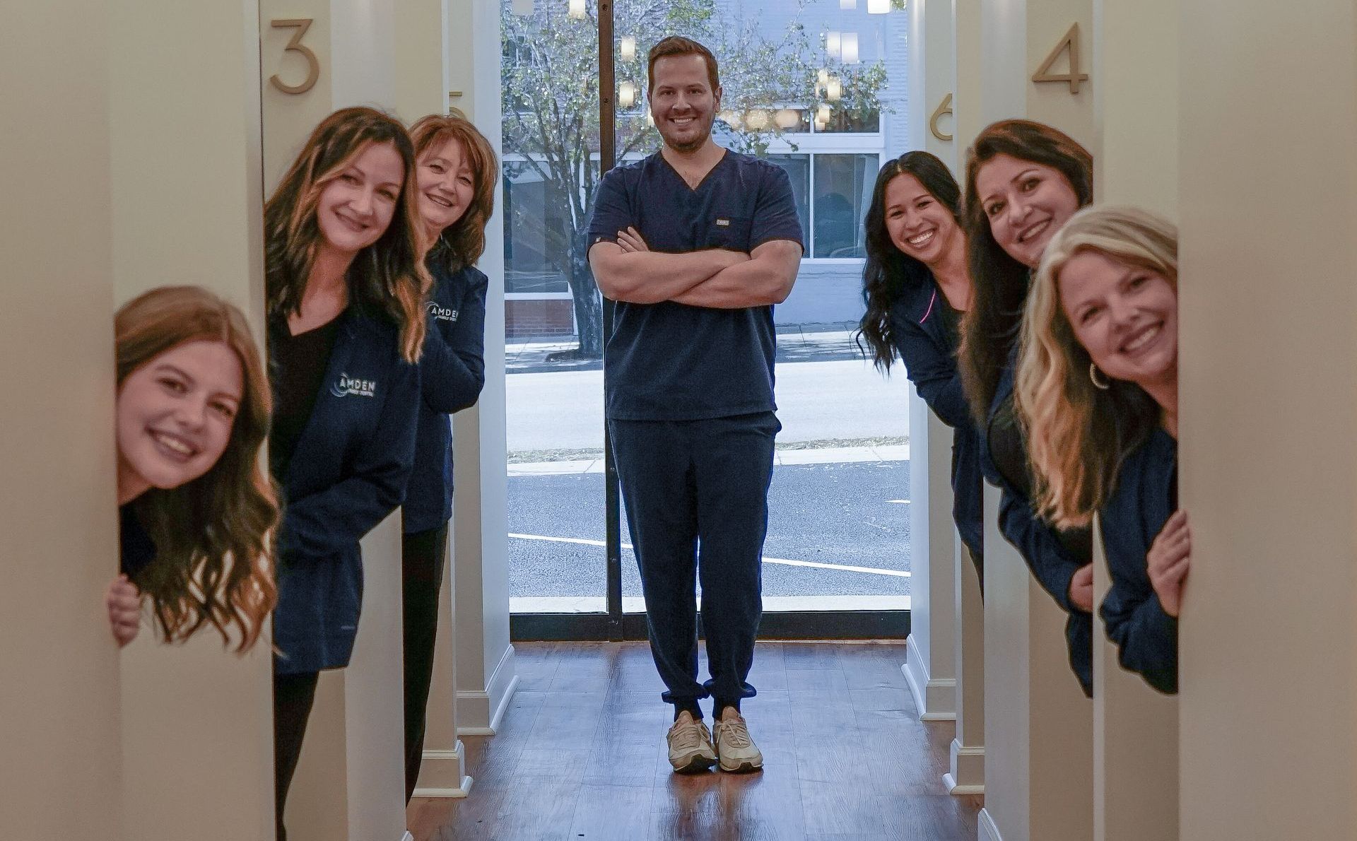 Dentist and staff smile, peering from doorways; sunny clinic with outdoor view.