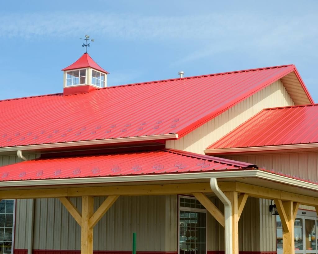 Red metal roof on a light-colored building with a cupola and a wooden porch under a blue sky.