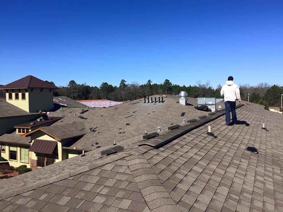 A person on a brown shingle roof inspects damage under a clear blue sky.