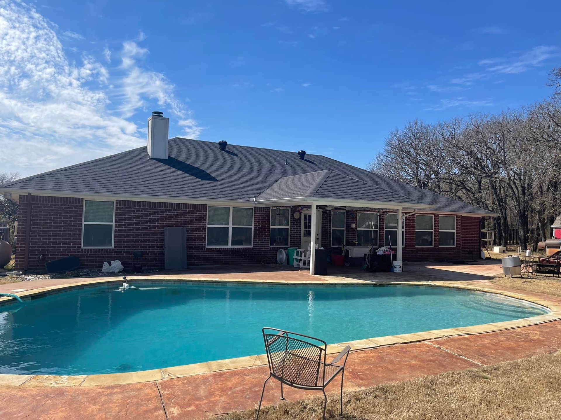 Backyard with a pool and red-brick house under a bright blue sky; a chair sits near the pool.