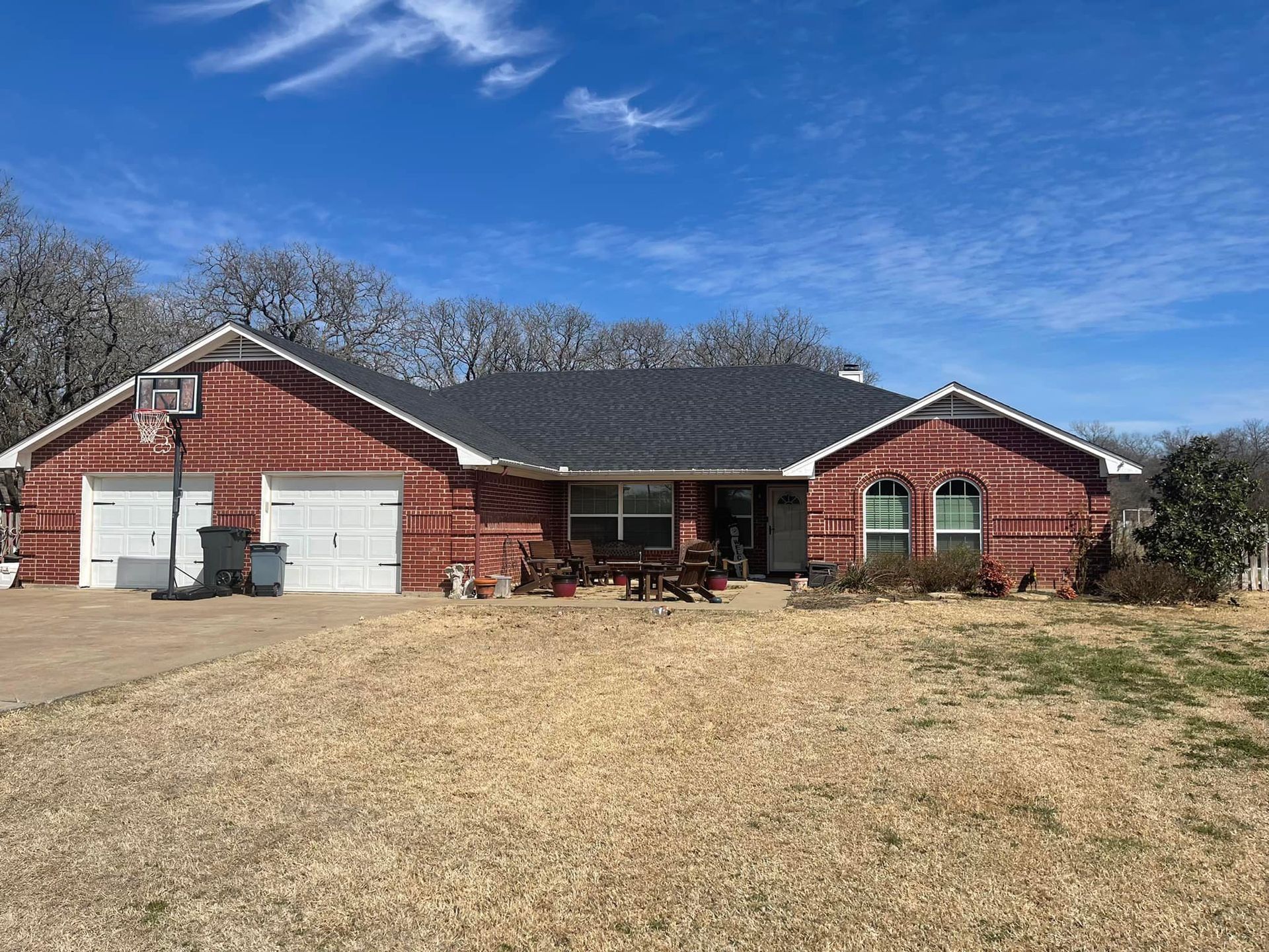 Red brick house with a two-car garage, blue sky, and brown yard. Basketball hoop visible.