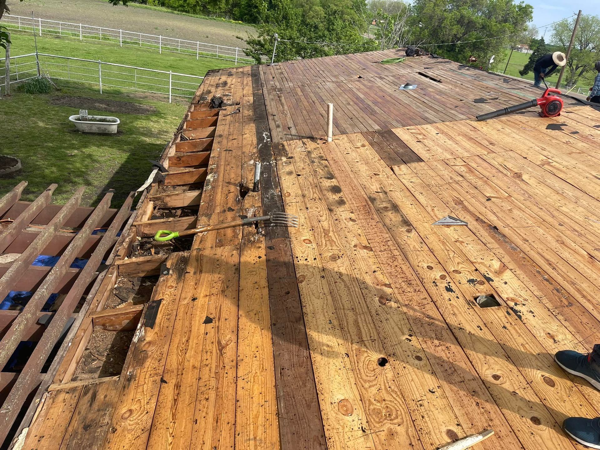 Roof being repaired, with old wooden boards exposed. Workers on roof, tools visible, green grass in background.
