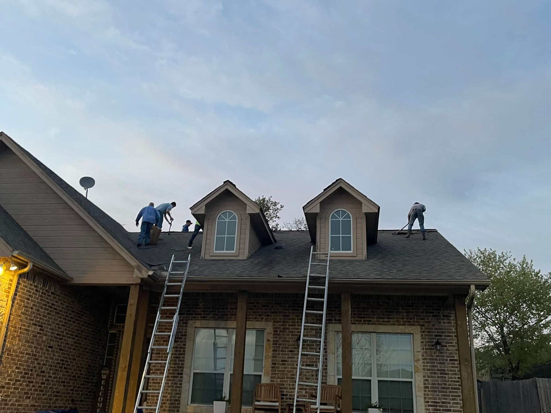Roofers on a brick house replacing shingles, using ladders under a cloudy sky.