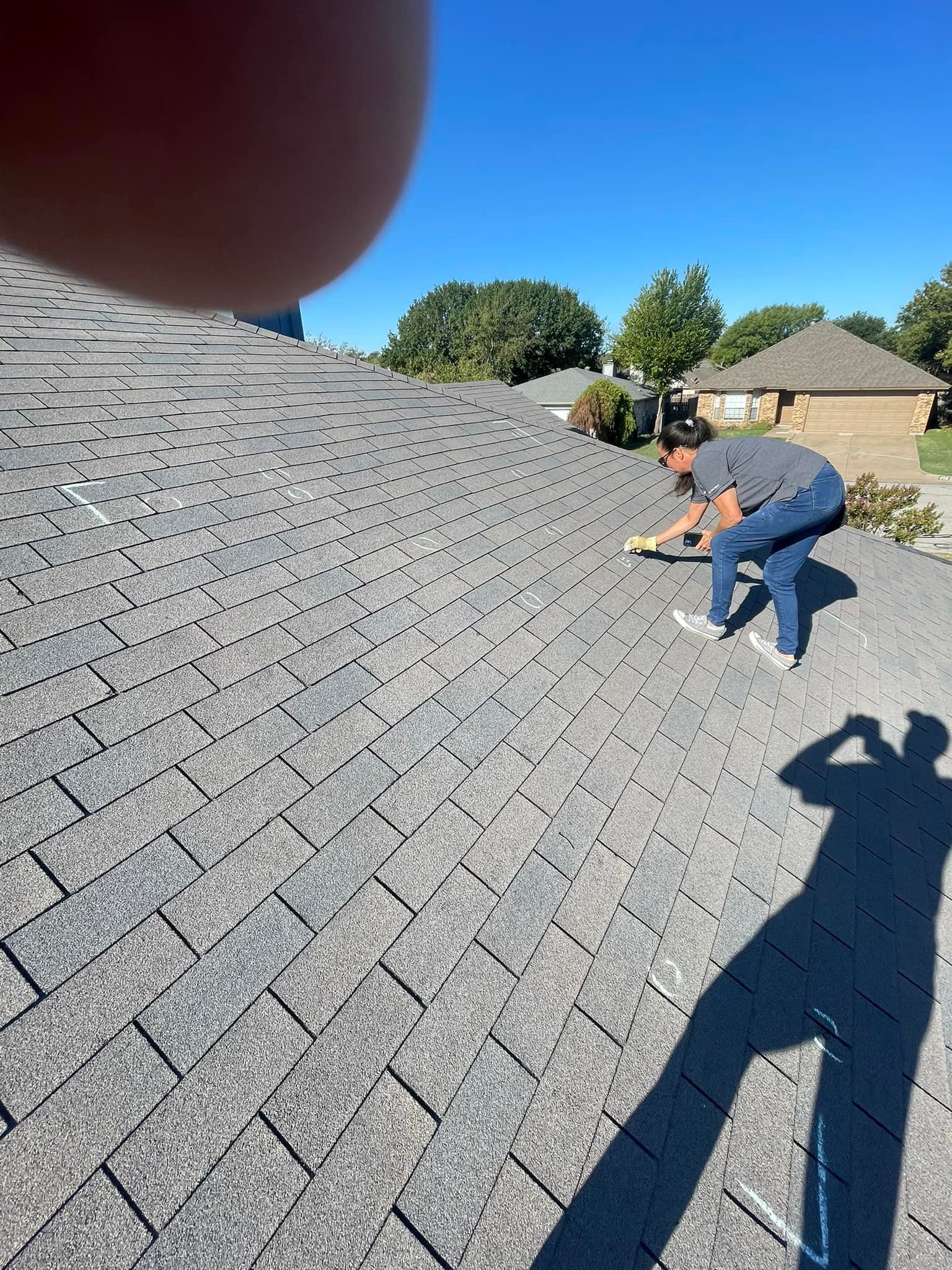 Person on a gray shingle roof, applying sealant on a sunny day. Their shadow and a blue sky are visible.