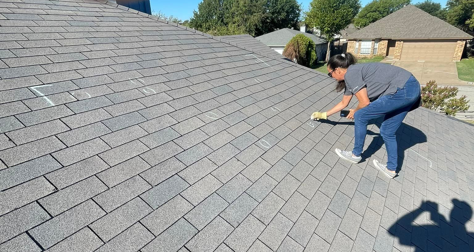 Woman on a gray shingle roof repairs shingles on a sunny day.