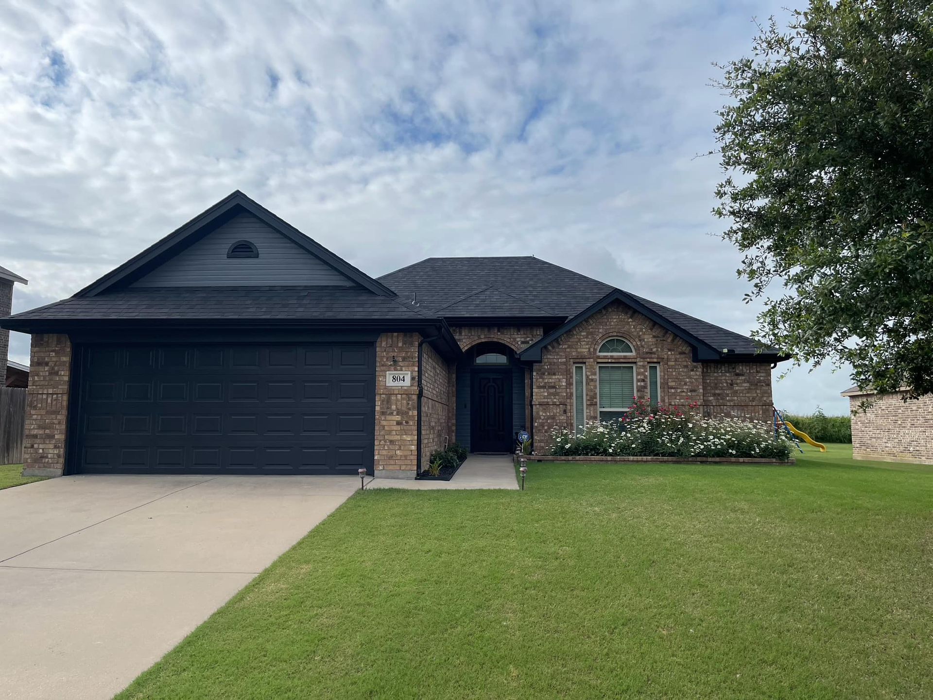 A single-story brick house with a black roof and garage door, green lawn, and cloudy sky.