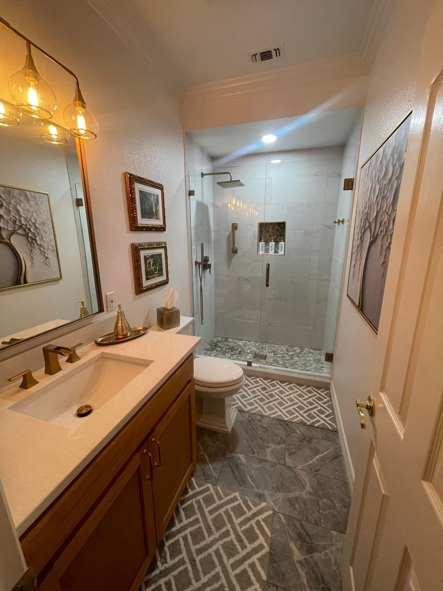 Bathroom with vanity, shower, toilet, and art. Wood vanity, gray tile floor, and light fixtures.