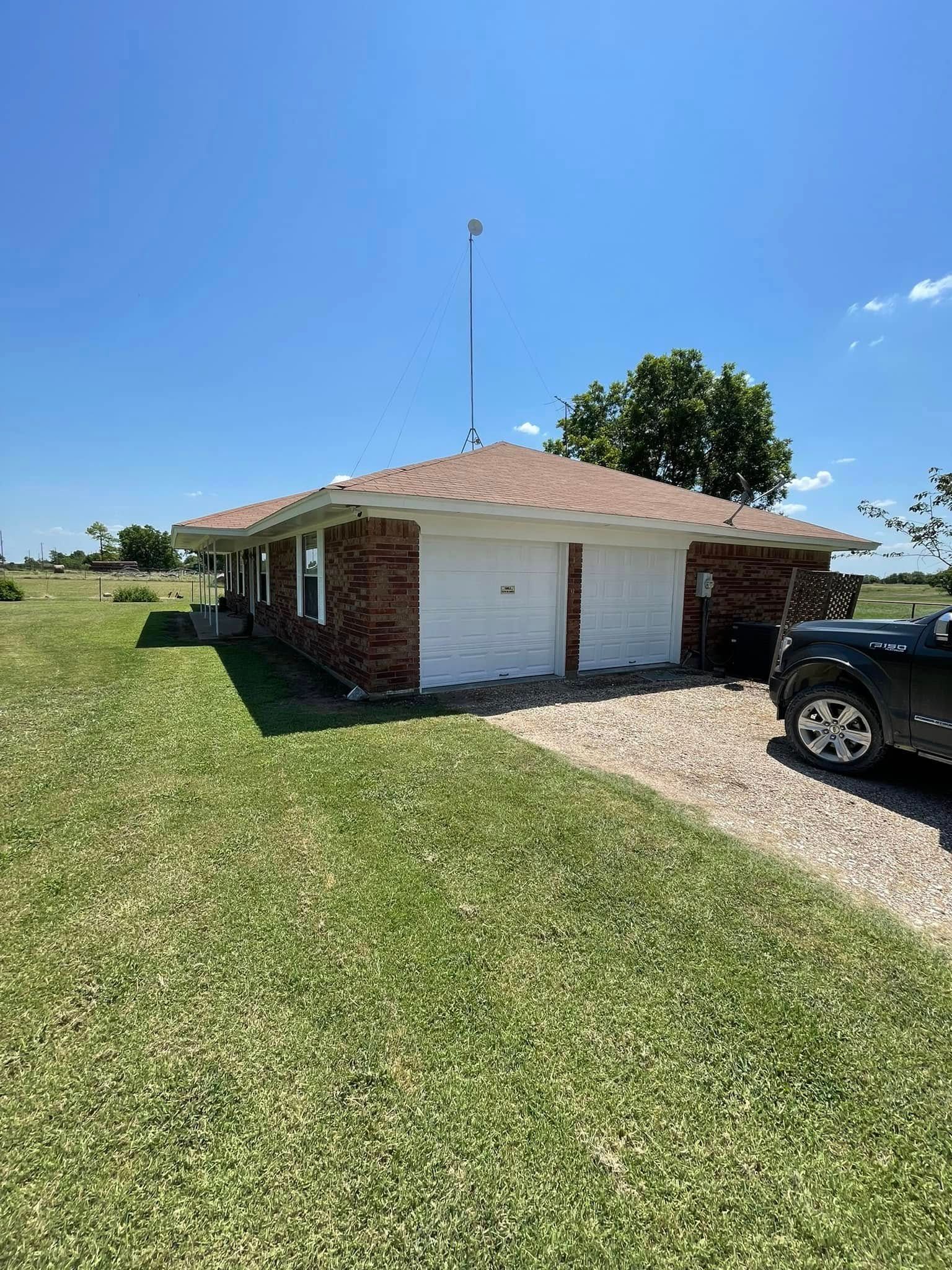 Brick house with two-car garage, gravel driveway, and green lawn under a blue sky. A black vehicle is parked outside.