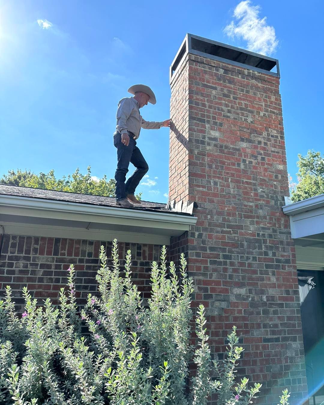Man in cowboy hat on a roof, touching a brick chimney, bright sky.
