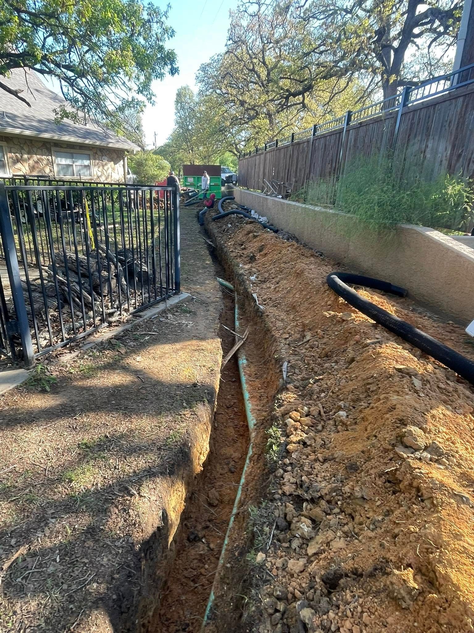 A trench dug alongside a fence with exposed pipes, possibly for utility work.