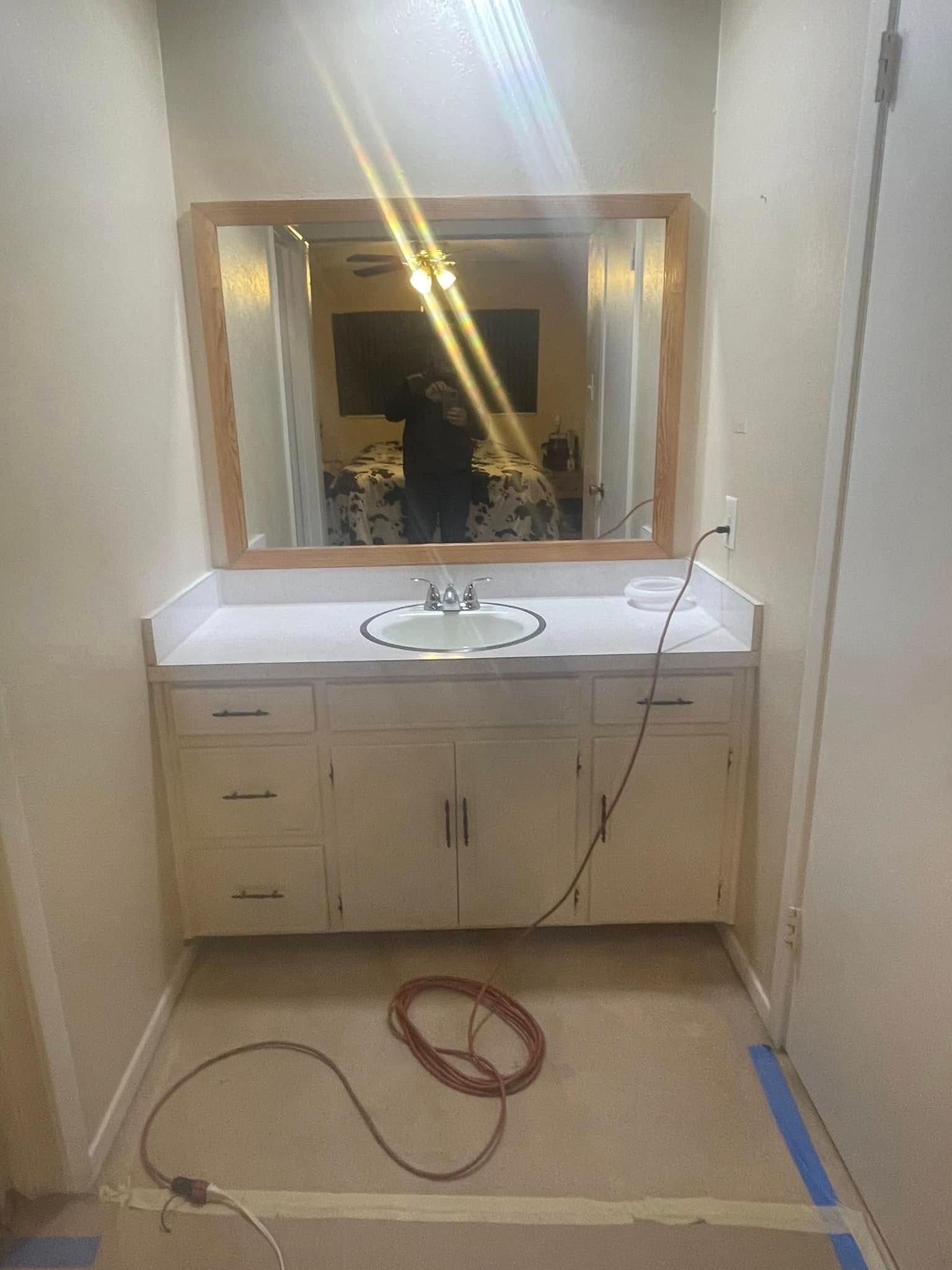 Bathroom with white vanity, light wood mirror frame, and an open door. Electrical cord on floor.