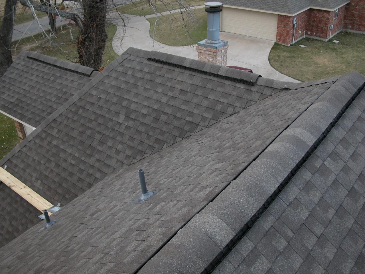Dark gray shingle roof with ridge cap, chimney, and visible vents on a house exterior.