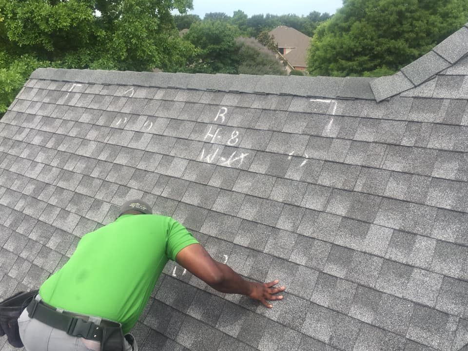 Person in green shirt inspecting a gray shingled roof, trees and a house in the background.