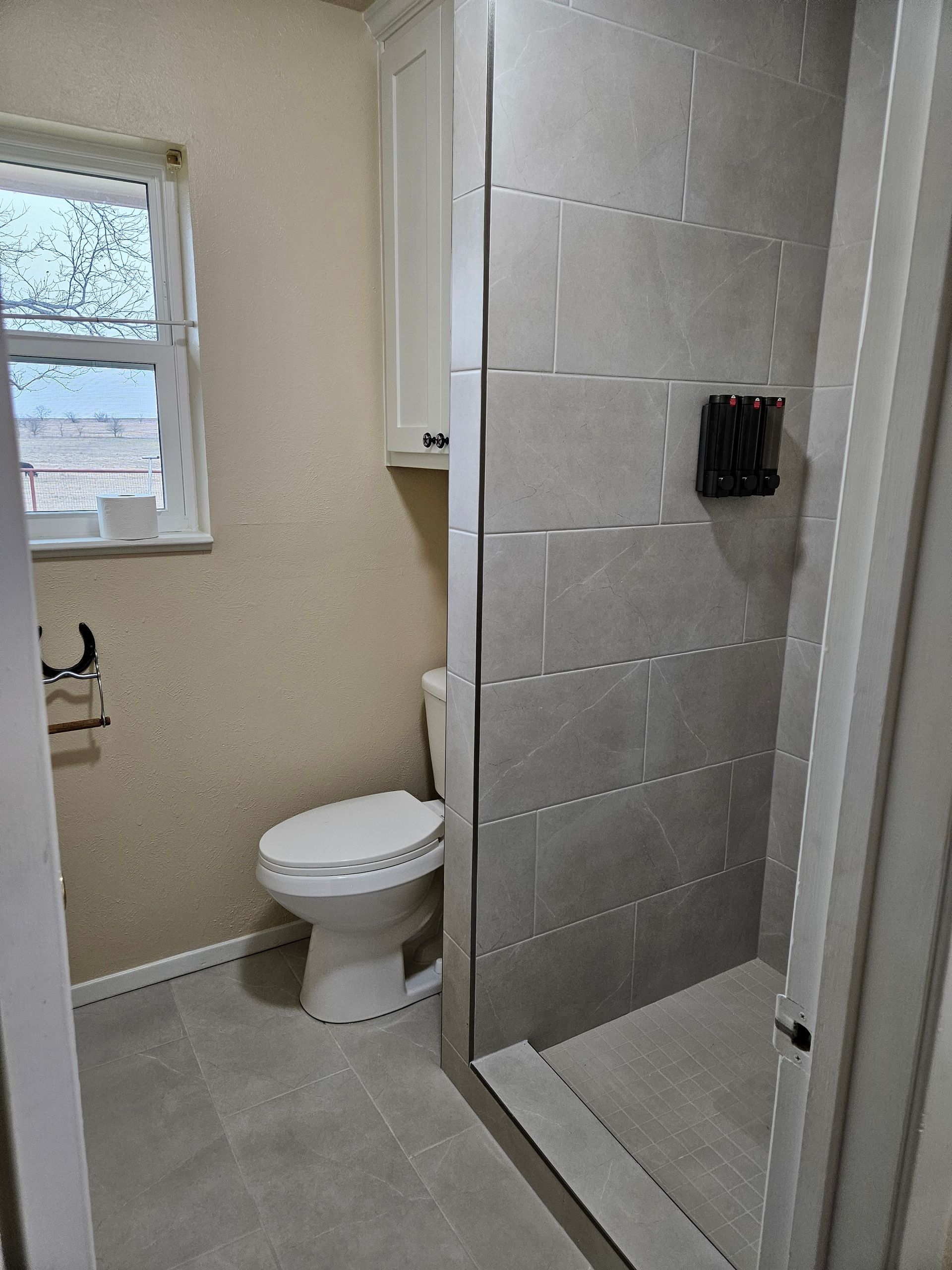 Bathroom with a toilet, shower, window, and cabinet; gray tiles and flooring.