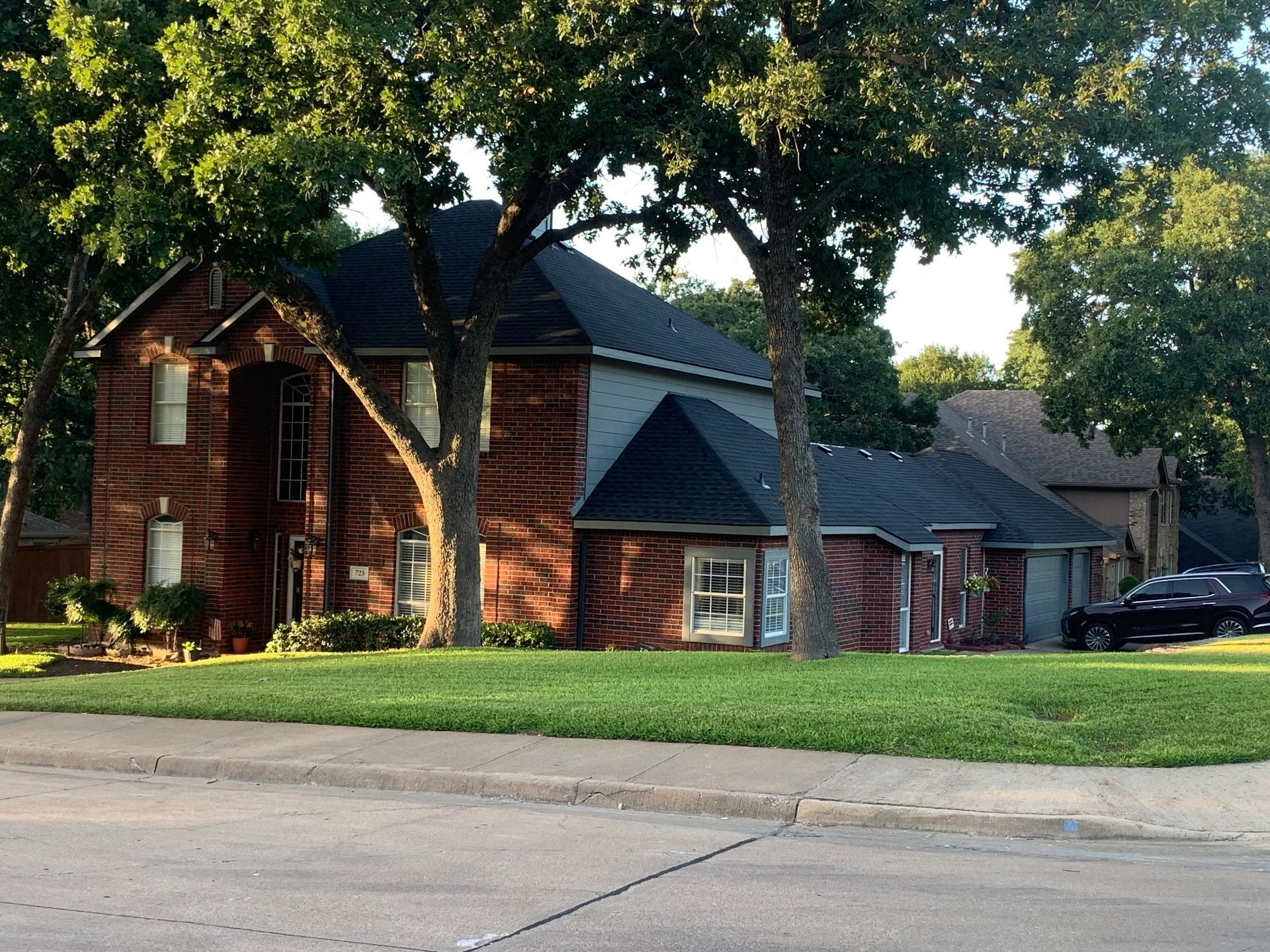 Two-story brick house with dark roof, green grass, and large trees on a corner lot.
