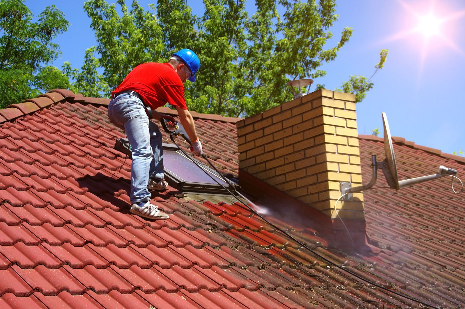 Man in red shirt and blue helmet pressure washing red tiled roof, near a brick chimney, on a sunny day.