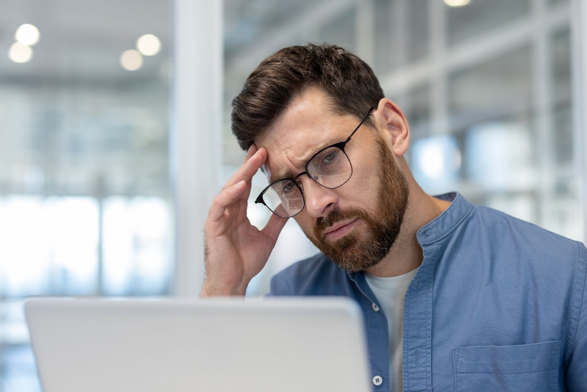Man with glasses, hand on forehead, looking stressed at a laptop in an office.