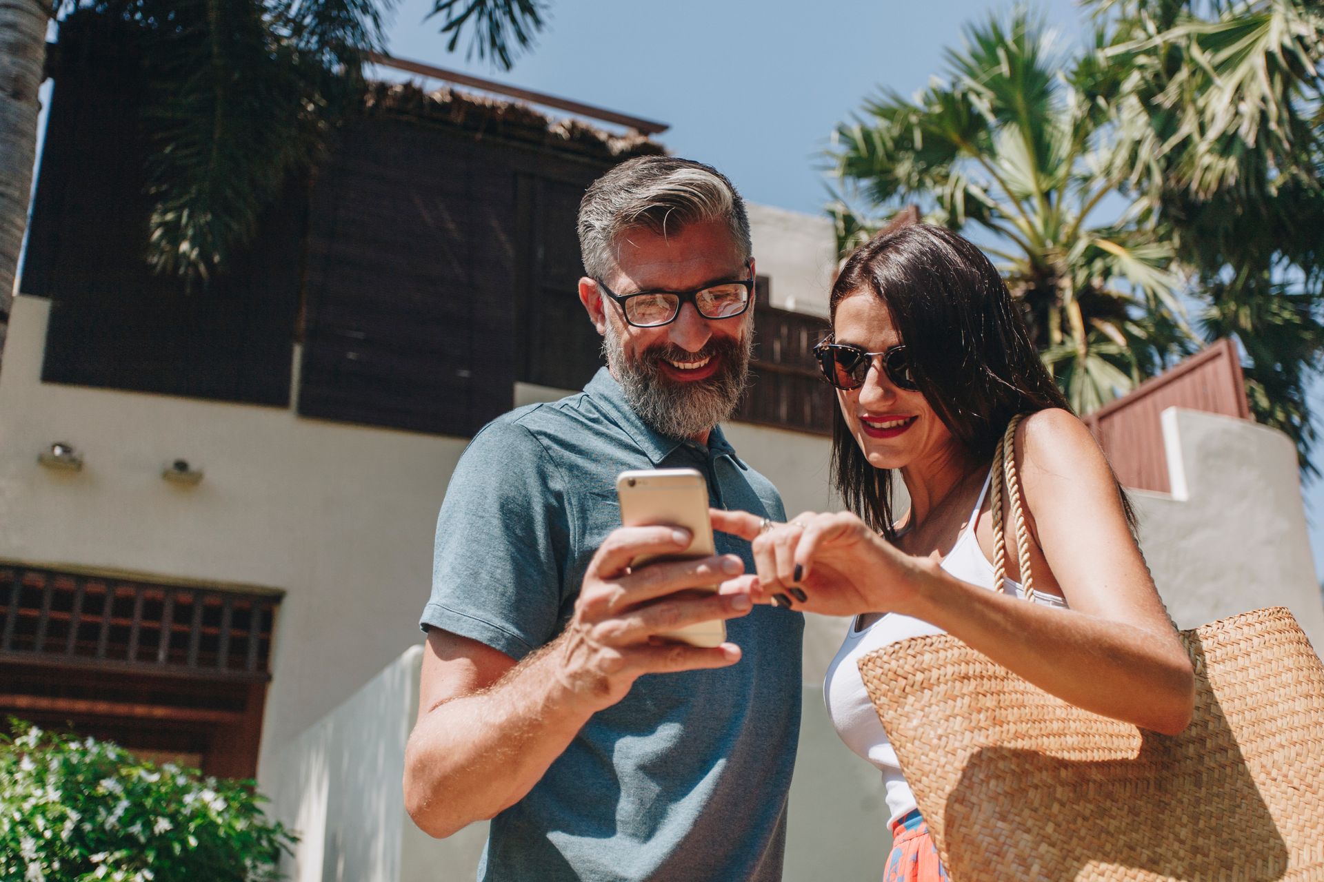 A couple looks at a smartphone together while standing outside a building on a sunny day.