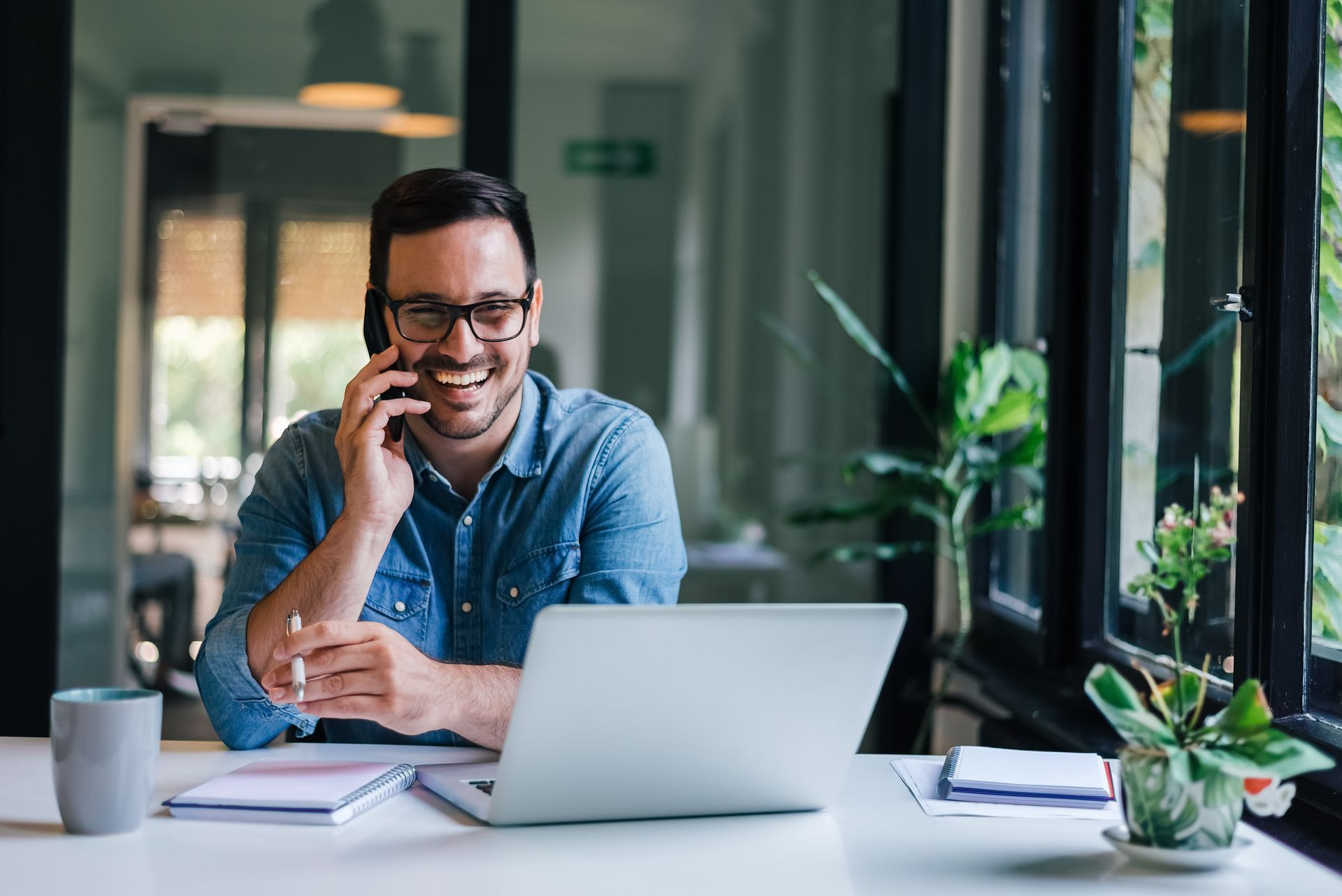 A smiling person in a denim shirt sits at a desk, talking on a phone while working on a laptop in a bright office.
