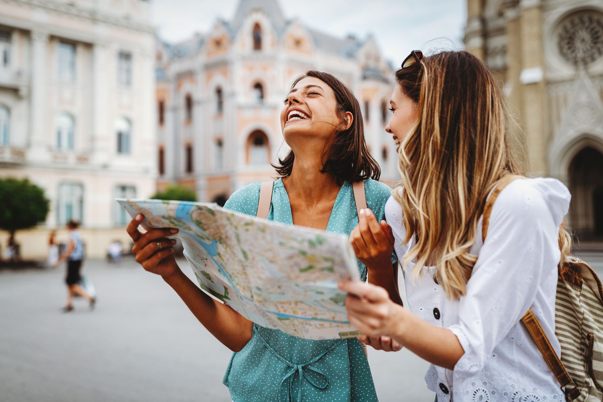 Two friends stand outdoors in a European city plaza, smiling and looking up while holding a large paper map.