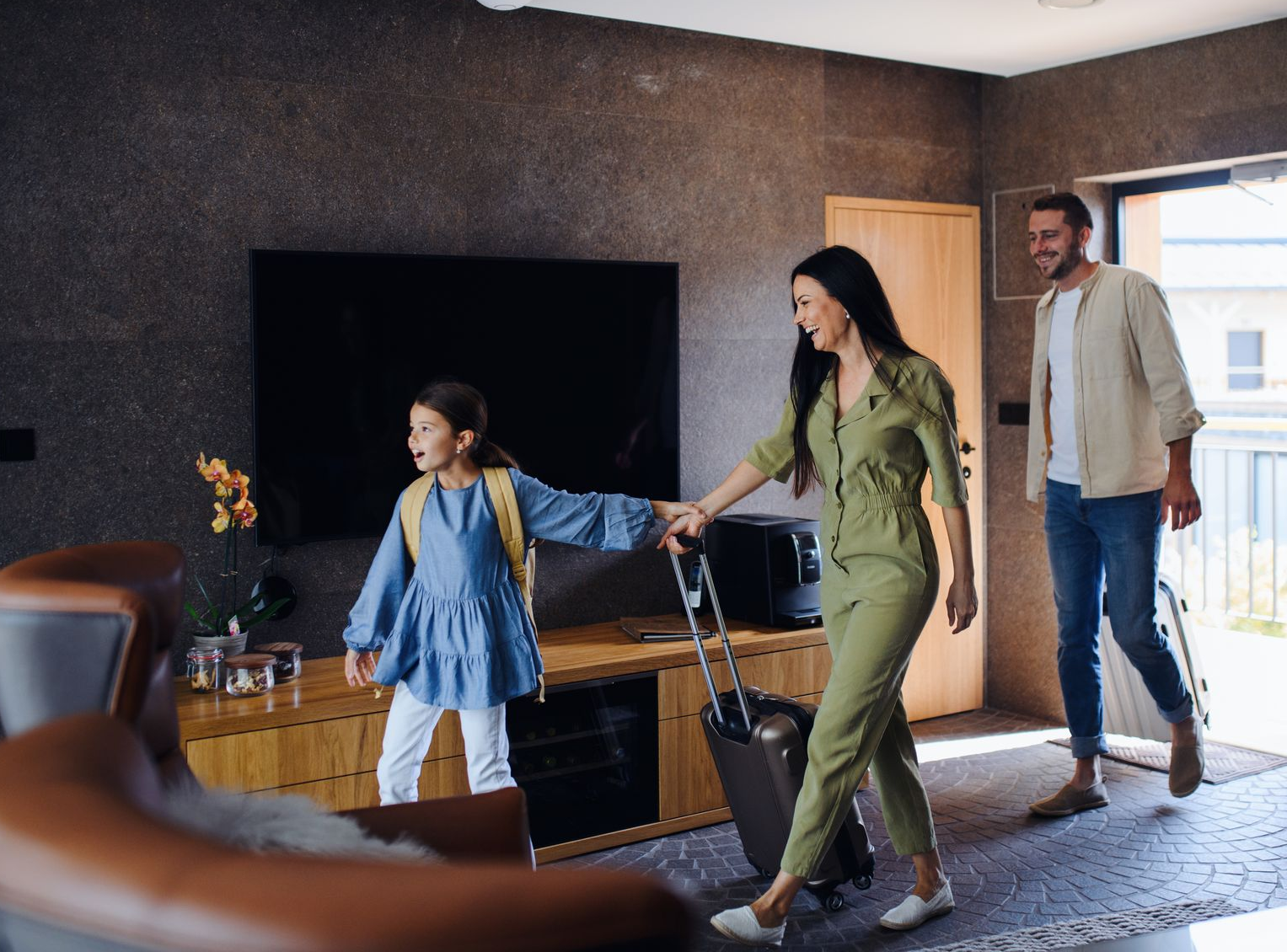 A family enters a modern living room carrying luggage, with a child in blue leading the way.