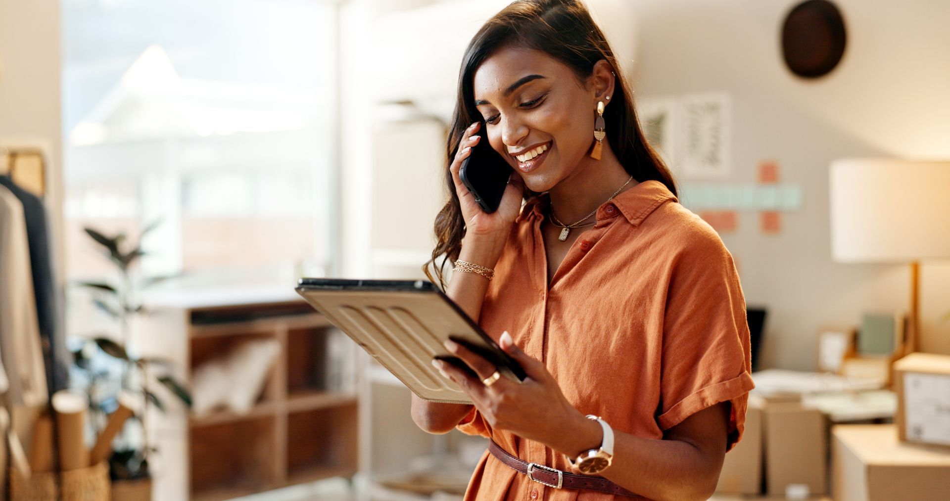 Smiling woman in an orange shirt talking on a phone while holding a tablet in a bright home office.