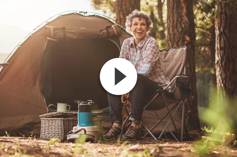 An elderly woman is sitting in a chair in front of a tent in the woods.