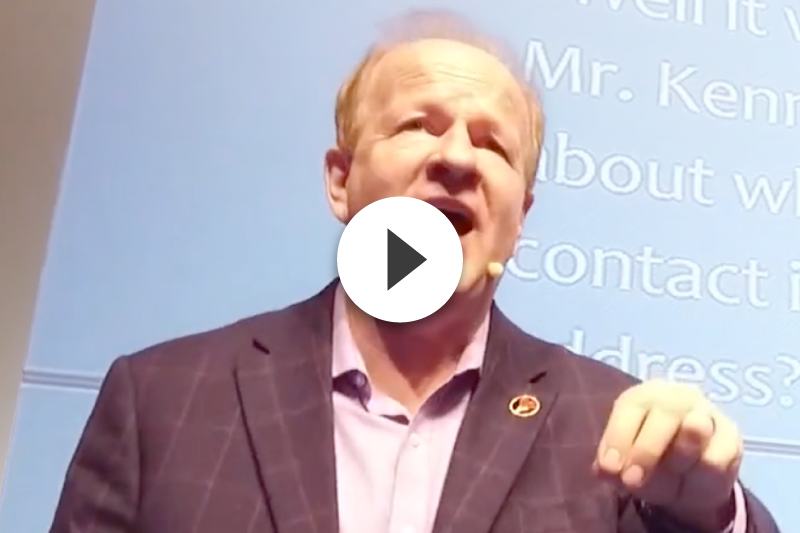 A man in a suit is giving a speech with a play button in front of him