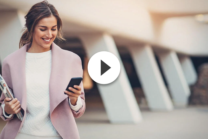 A woman in a pink coat is walking down the street while looking at her cell phone.