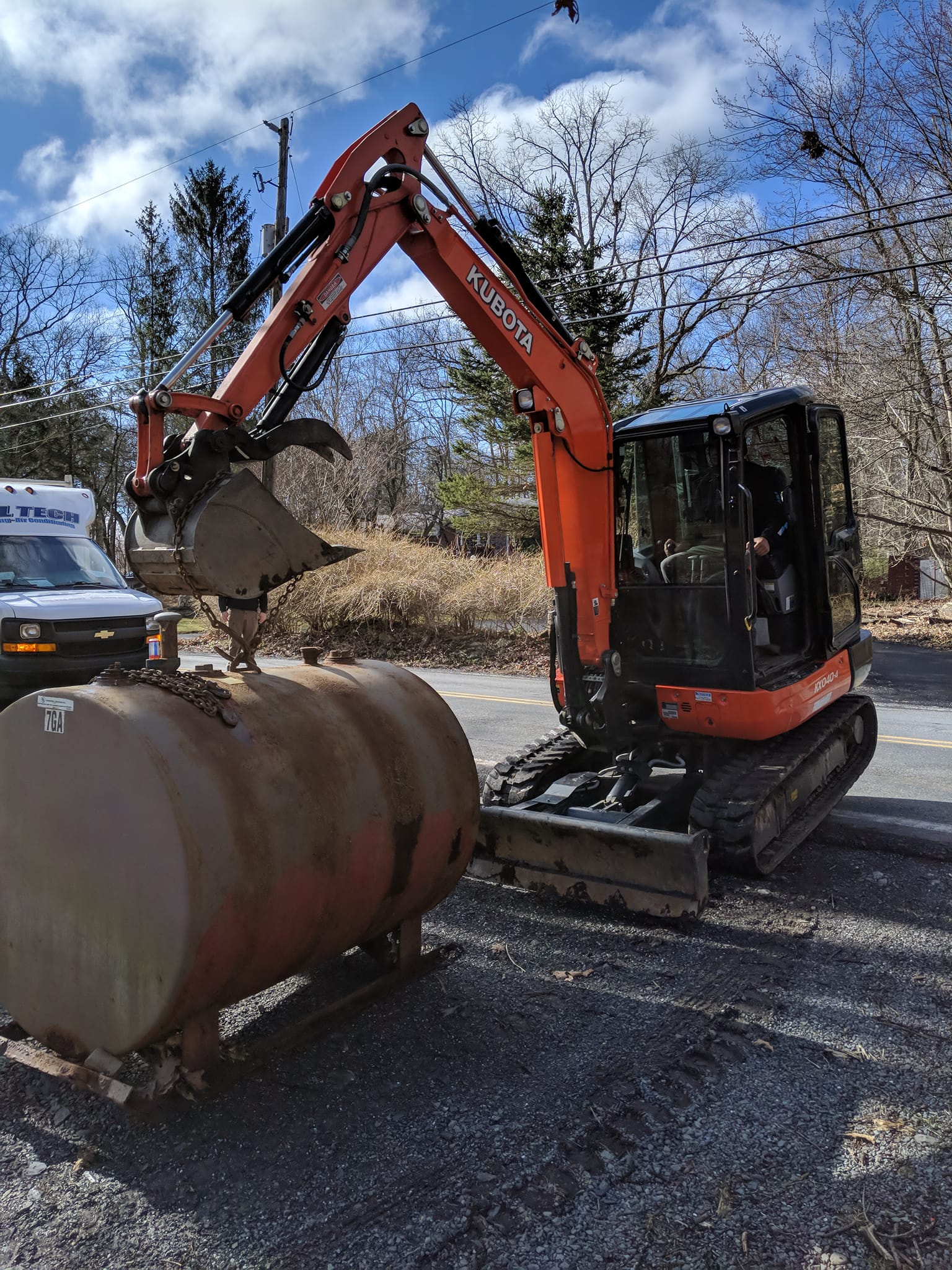 A small orange excavator is loading a large tank on the side of the road.