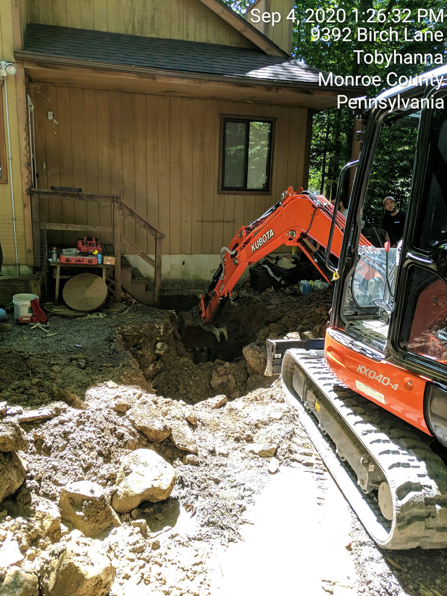 A small orange excavator is digging a hole in the dirt in front of a wooden house.